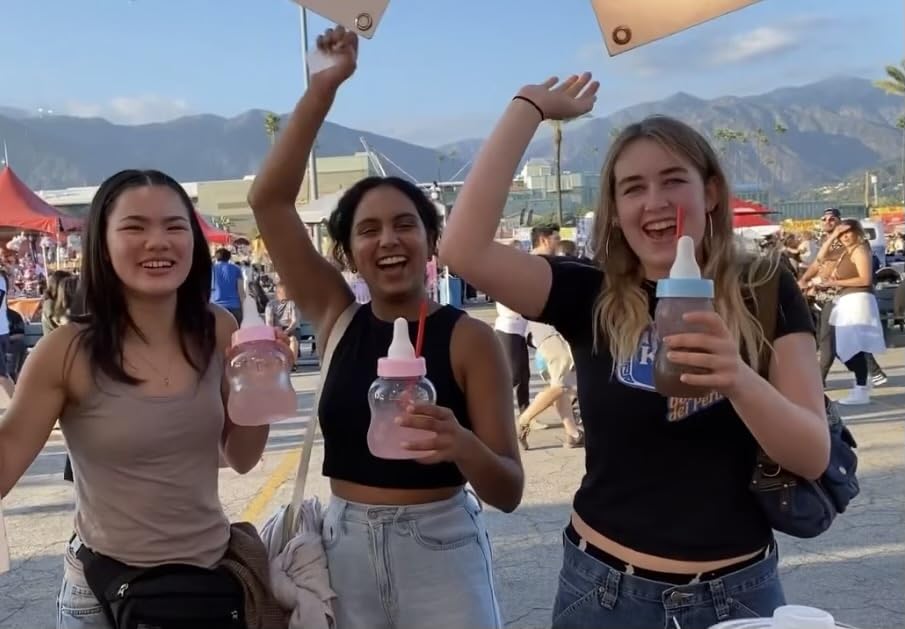 Three women holding piggy bank bottles with pink lemonade in a lively outdoor setting with mountains in the background.